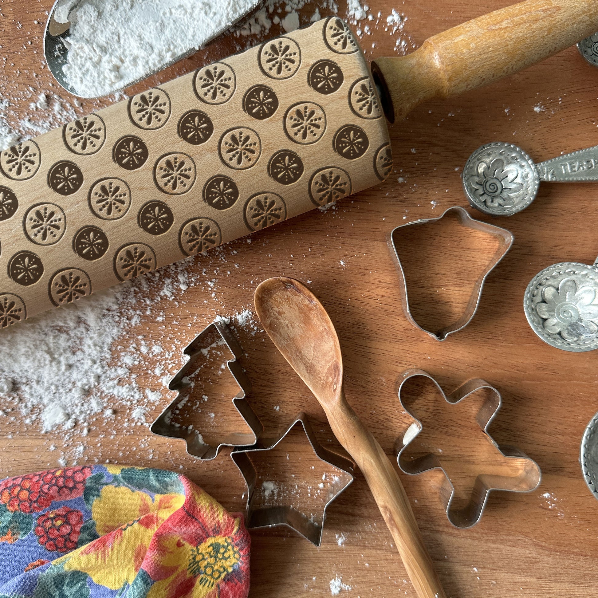 Baking tools including a rolling pin with embossed designs, cookie cutters, and a wooden spoon on a wooden surface.