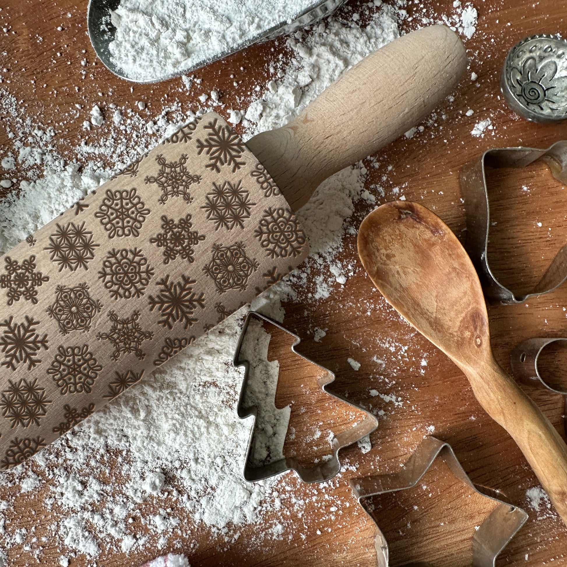 Wooden rolling pin with snowflake patterns on a flour-dusted surface with cookie cutters.