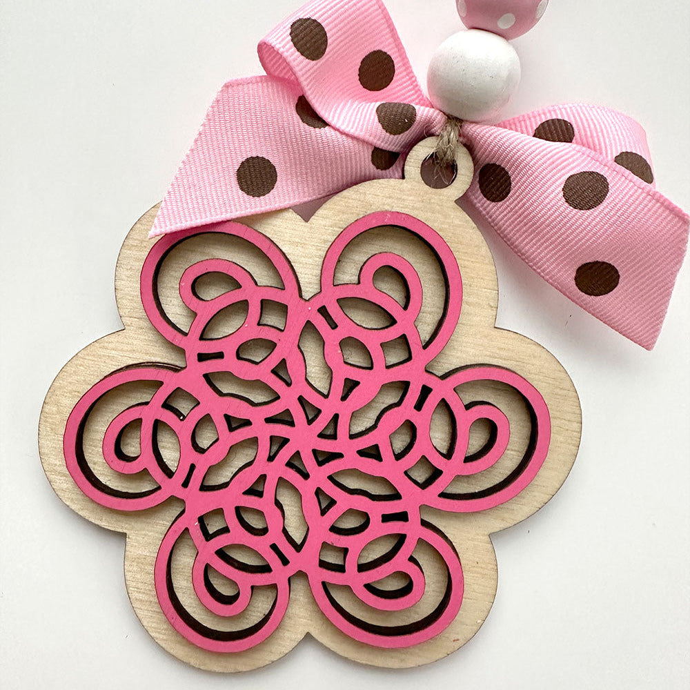 Decorative wooden wall hanging with pink intricate design and polka dot ribbon on a white background