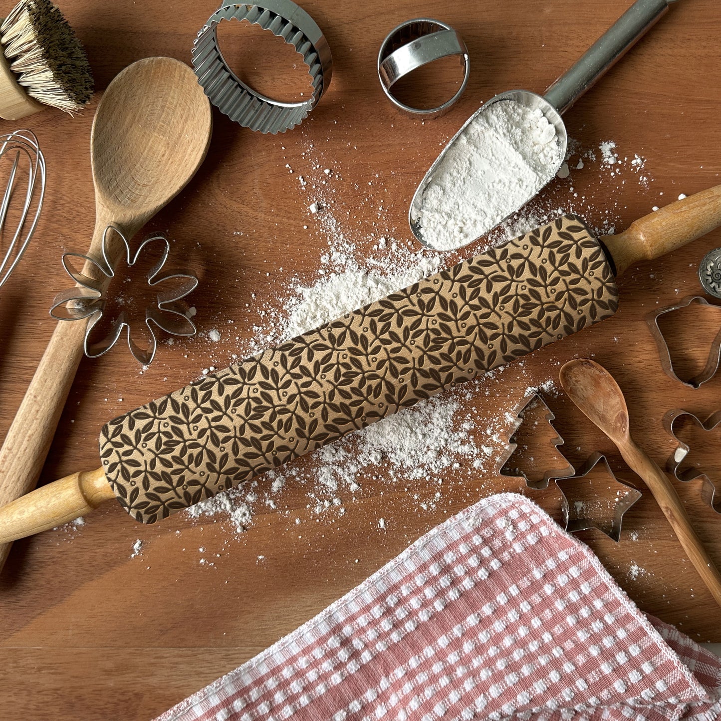 Baking tools including a rolling pin with a pattern, wooden spoon, and cookie cutters on a wooden surface with flour.
