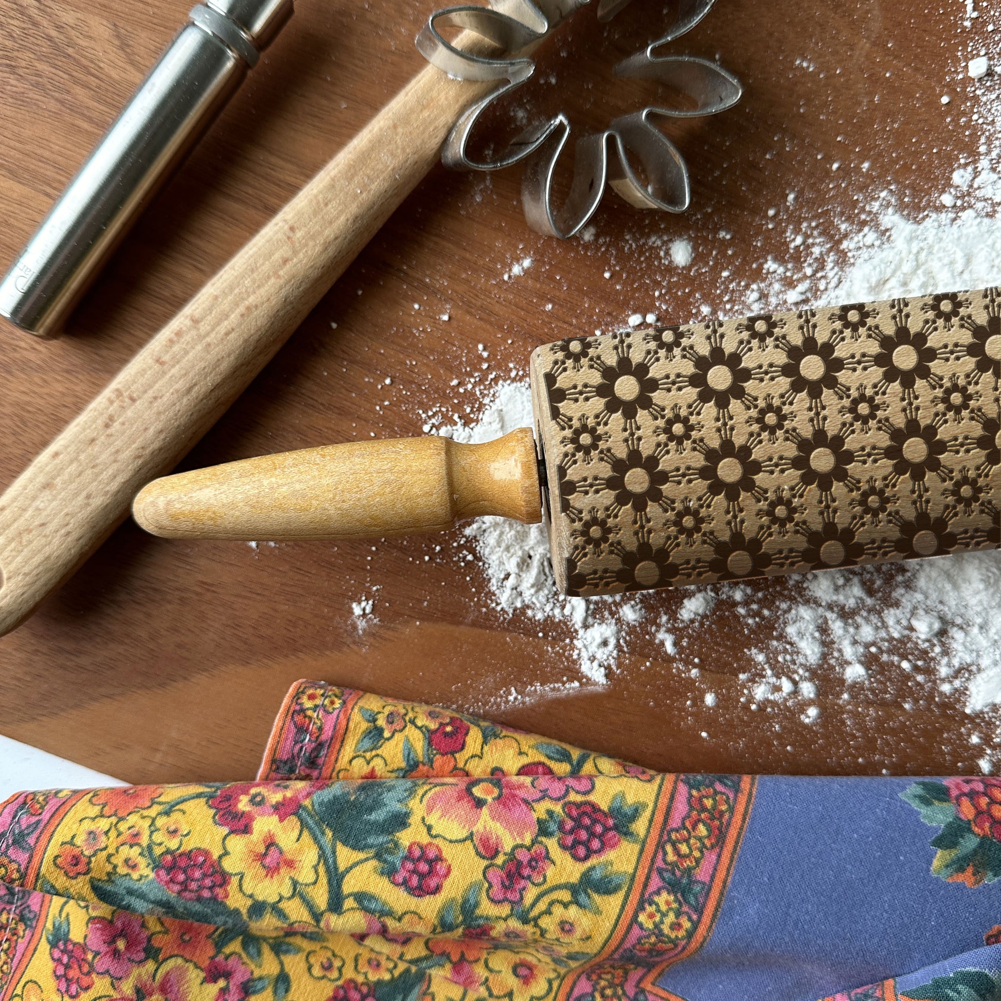 Decorative rolling pin with floral patterns on a wooden surface with flour and cookie cutters.
