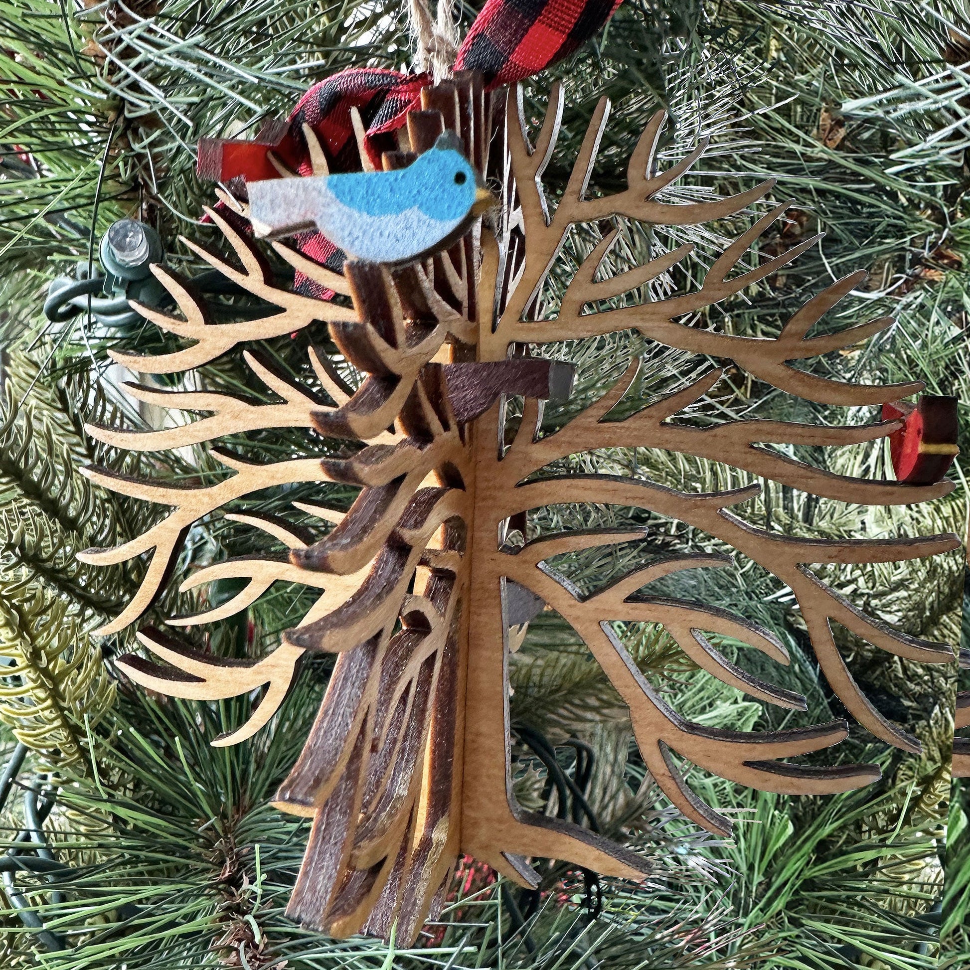 Decorative wooden ornament with a bird on a tree branch, hanging from a red and black plaid ribbon, against a Christmas tree background.
