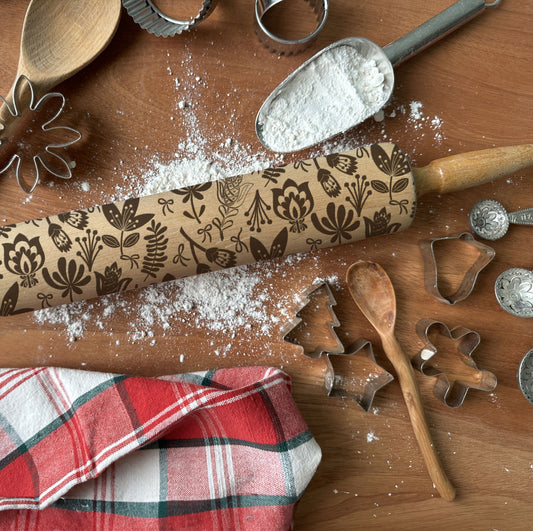 Wooden rolling pin with embossed patterns on a wooden surface with flour, cookie cutters, and a red checkered towel.