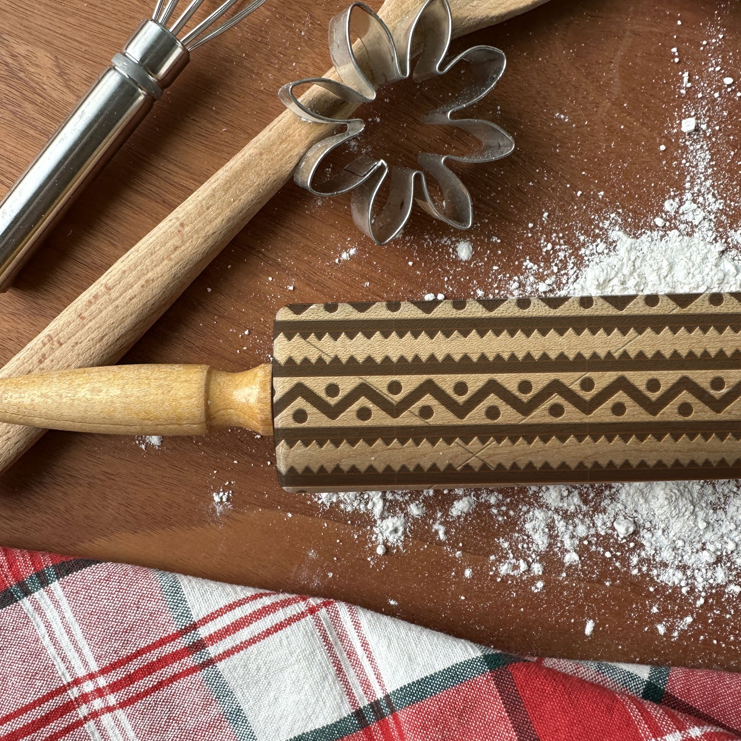 Decorative rolling pin with pattern, whisk, and flower cookie cutter on a wooden surface with flour.